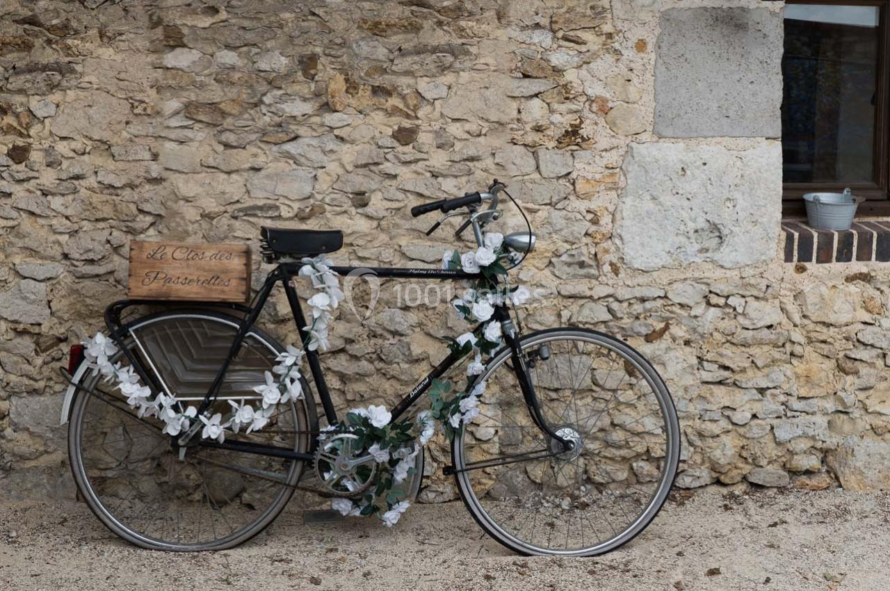 Vélo noir décoré de fleurs blanches et vertes, appuyé contre un mur en pierre devant une fenêtre.