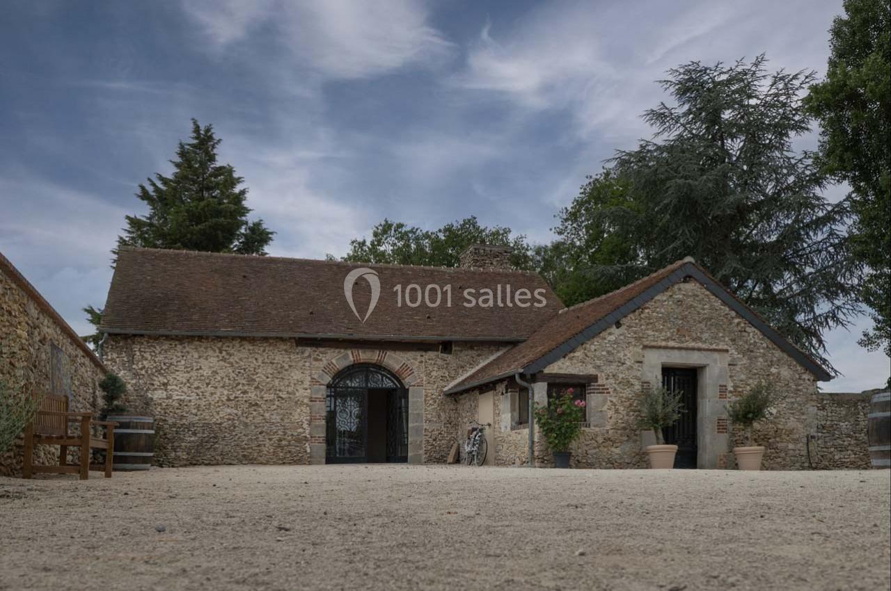 Bâtiment en pierre avec toit en tuiles, entouré d'arbres, sur une cour en gravier sous un ciel partiellement nuageux.