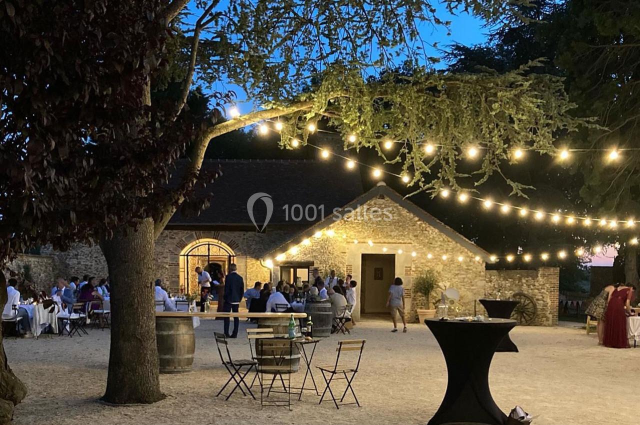 Cour extérieure éclairée par des guirlandes lumineuses, avec des tables et des invités rassemblés près d'un bâtiment en…