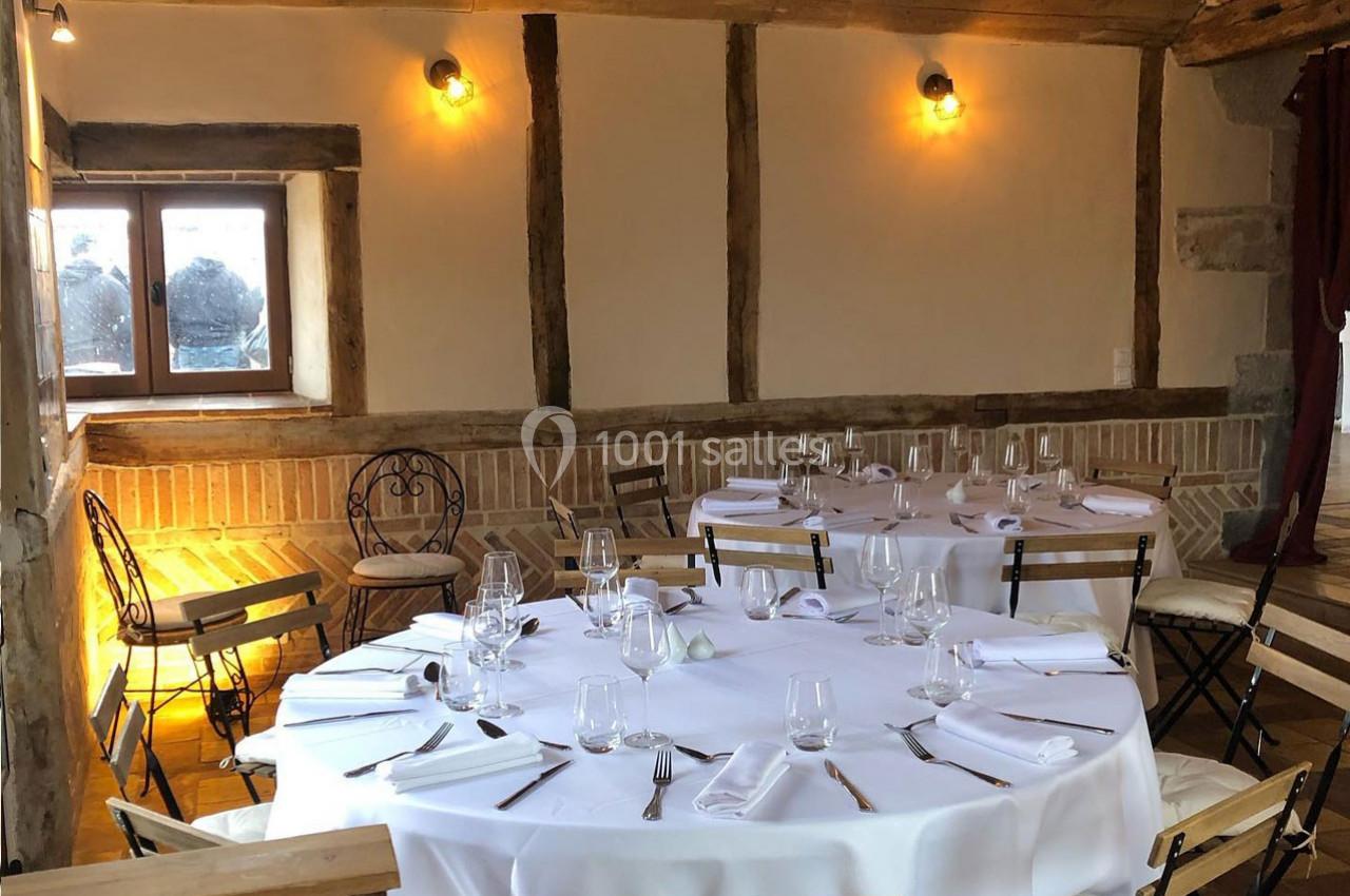 Salle rustique avec murs en bois et briques, tables rondes dressées avec nappes blanches et chaises en métal.