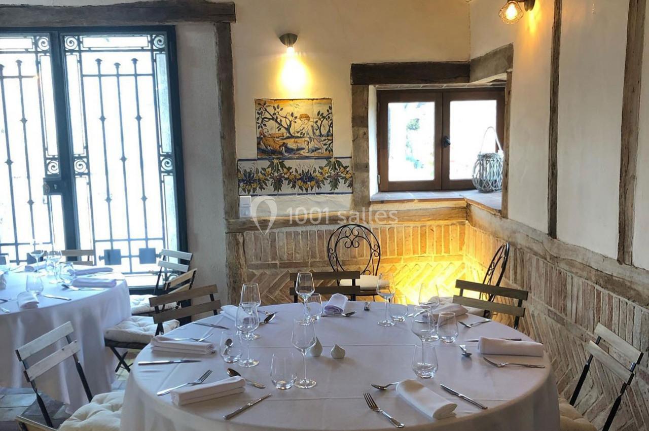 Salle de restaurant rustique avec tables rondes dressées, murs en bois et briques, et lumière naturelle.