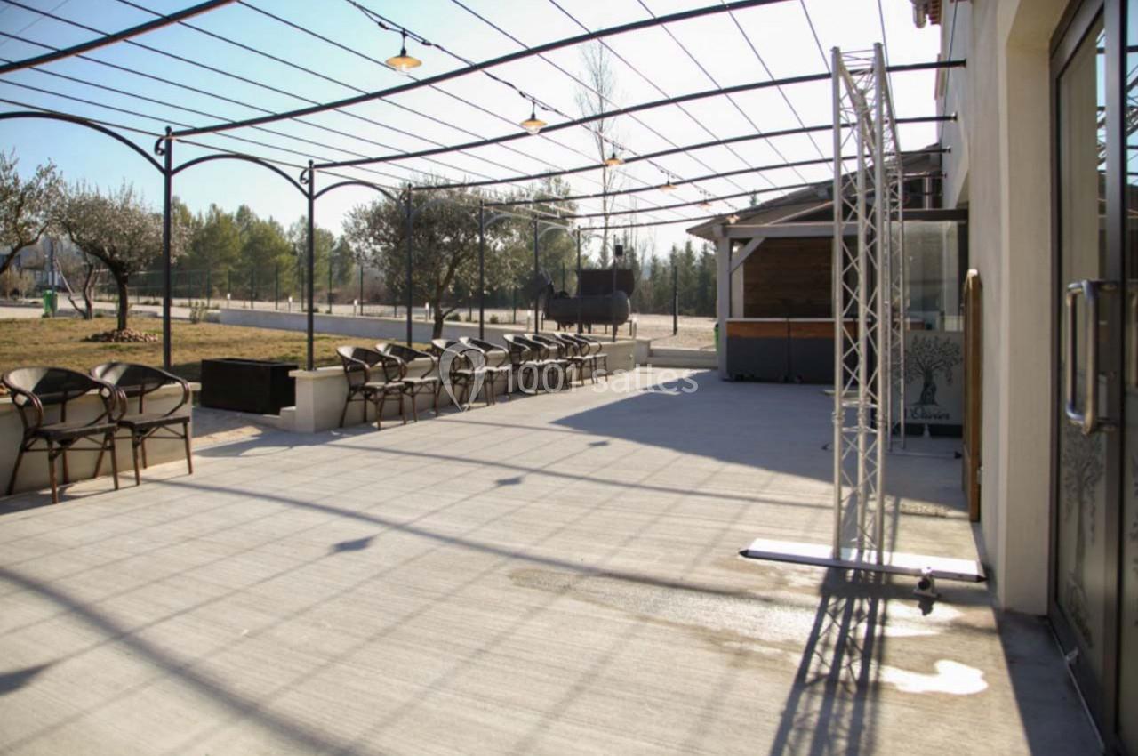 Terrasse extérieure avec pergola métallique, chaises alignées, arbres en arrière-plan et sol en béton clair.