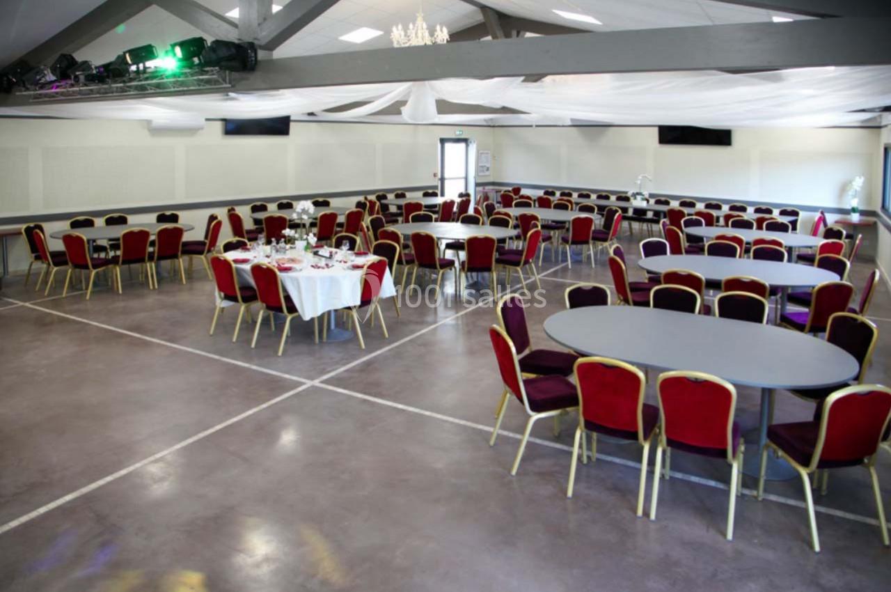 Salle de réception avec tables rondes et chaises rouges, décor sobre et plafond avec poutres apparentes.
