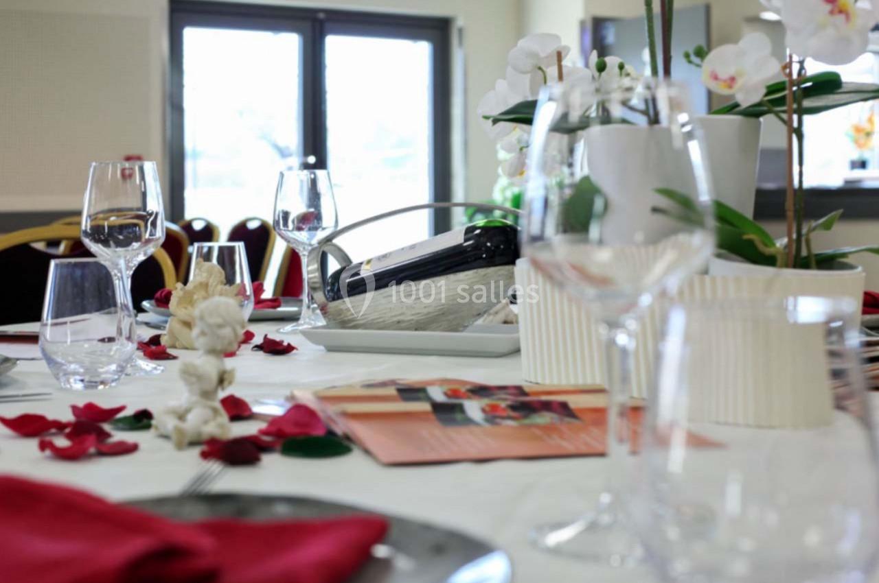 Table dressée avec nappes blanches, pétales rouges, verres, assiettes et décoration florale dans une salle lumineuse.
