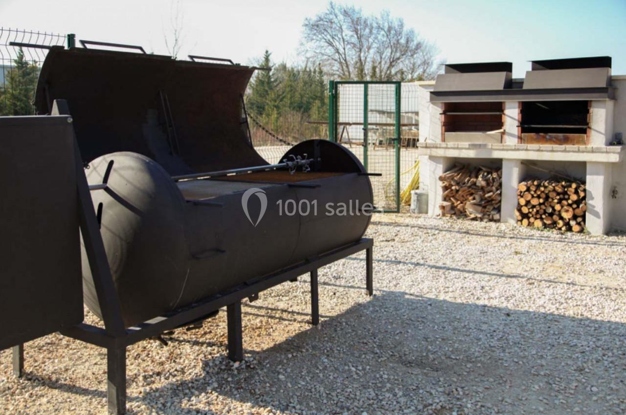 Barbecue fumoir en métal noir et four à bois en arrière-plan, entourés de graviers et de bois empilé.