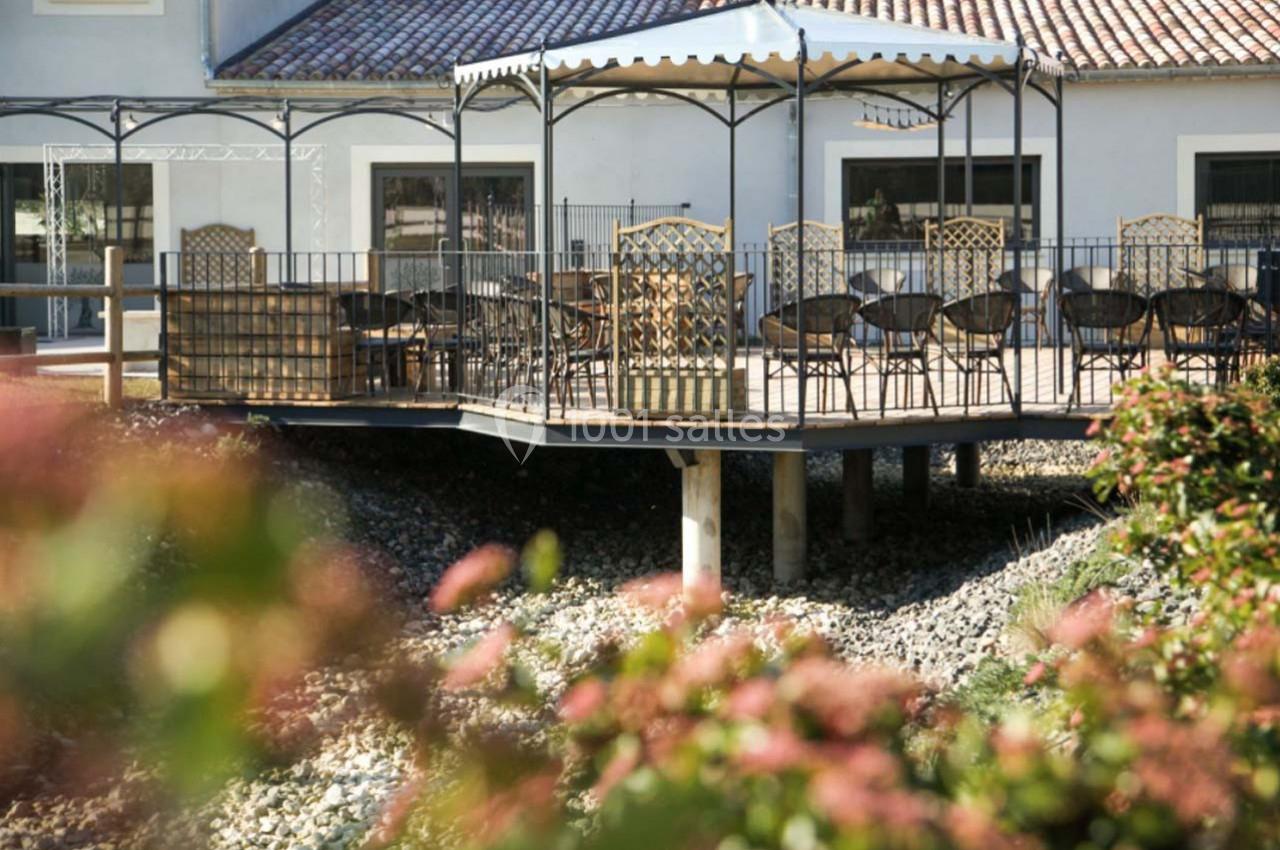 Terrasse surélevée avec pergola, équipée de tables et chaises, entourée de végétation et située devant un bâtiment clair.