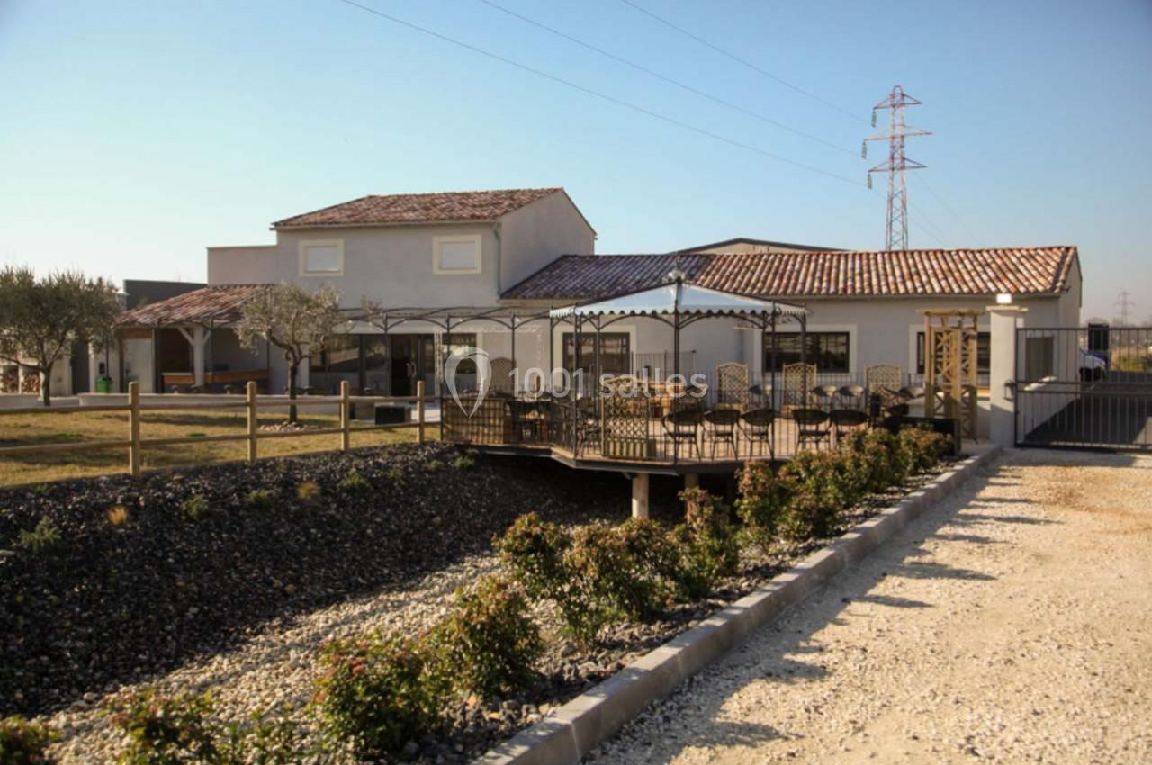 Maison avec terrasse en bois couverte, jardin paysager et allée gravillonnée, située dans un environnement rural.