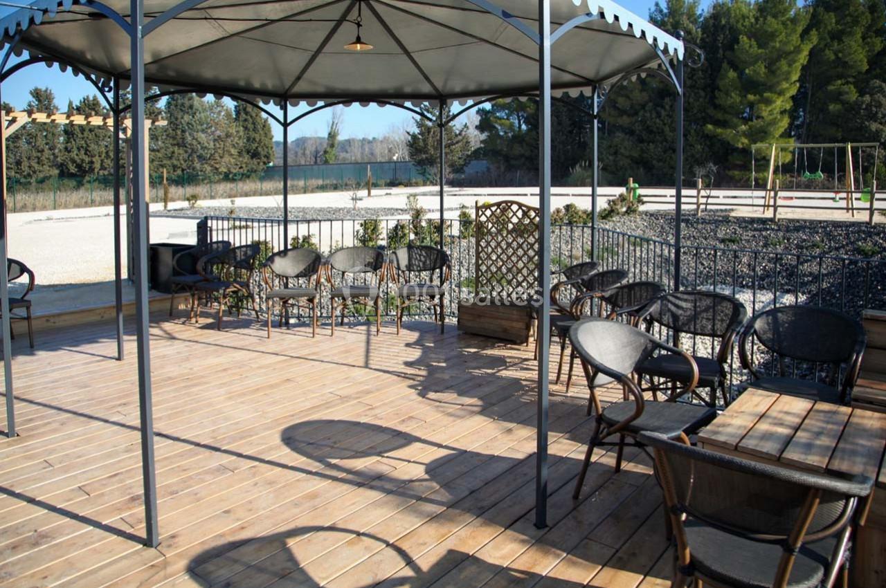 Terrasse en bois avec tables et chaises sous une pergola, entourée de végétation et d'une aire de jeux en arrière-plan.