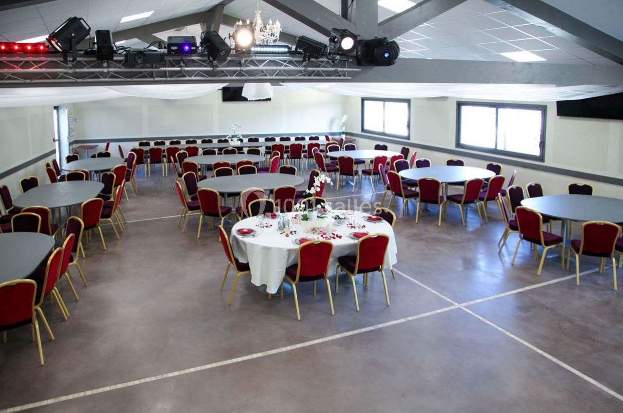 Salle de réception avec tables rondes dressées, chaises rouges et éclairage naturel provenant des fenêtres.