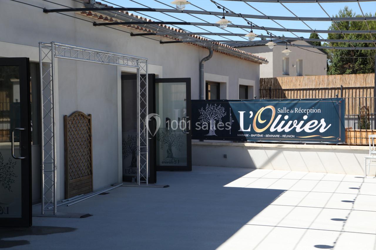 Entrée d'une salle de réception avec une bannière ’L'Olivier’ et des portes ouvertes donnant sur une terrasse.