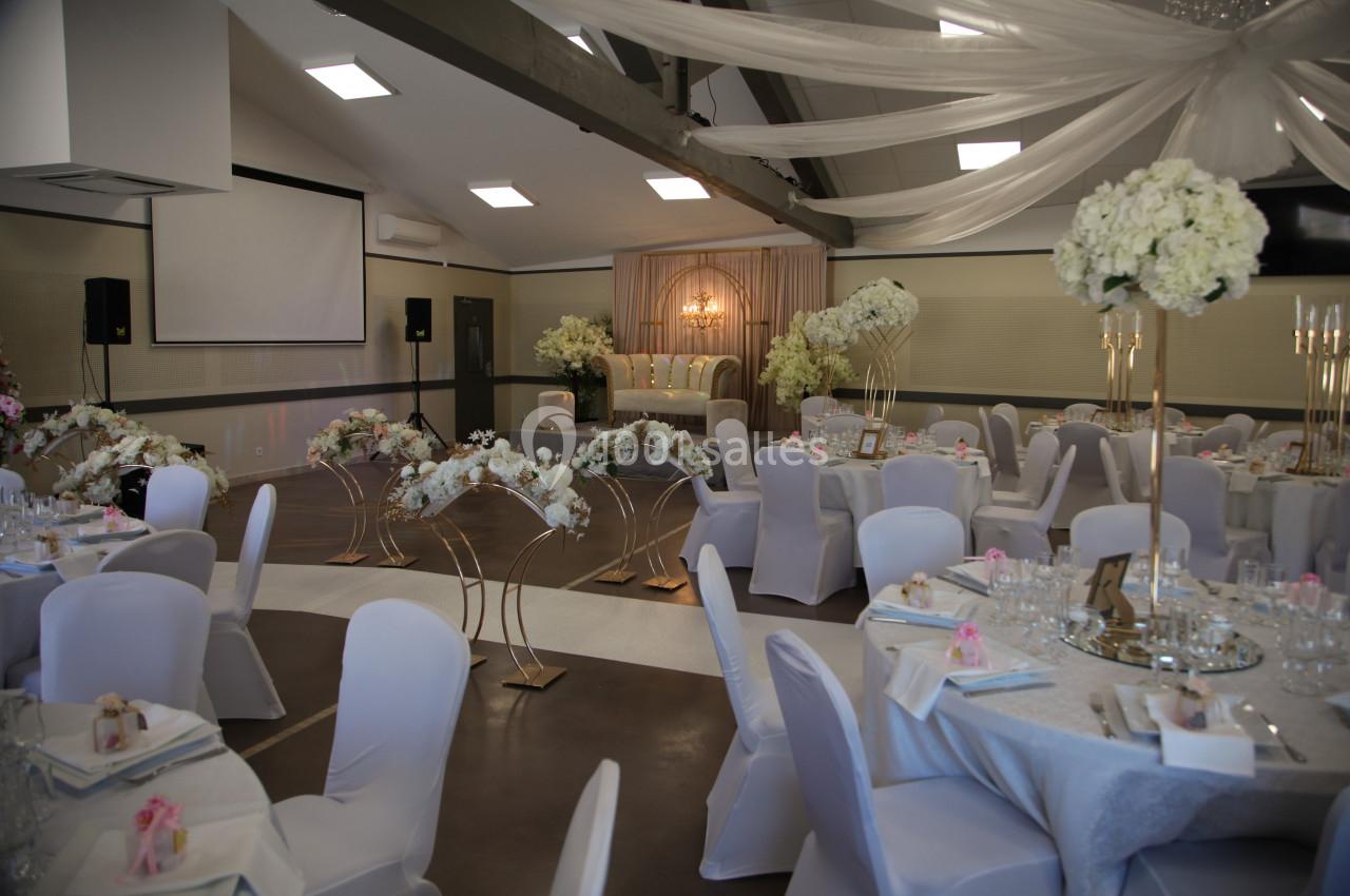 Salle de réception décorée avec des tables rondes, nappes blanches, compositions florales et un espace scénique éclairé.