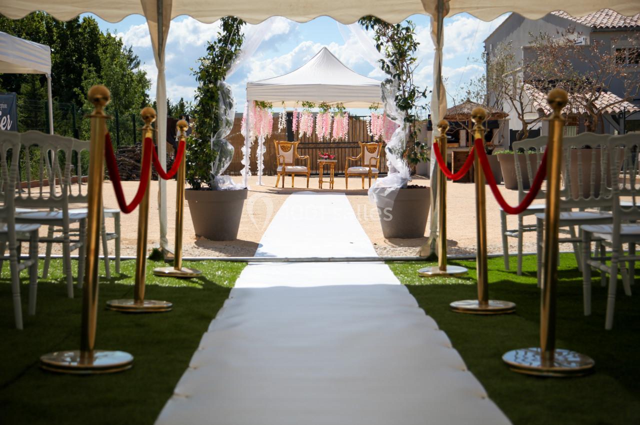 Allée centrale menant à une arche décorée pour une cérémonie en extérieur, entourée de chaises et de plantes.