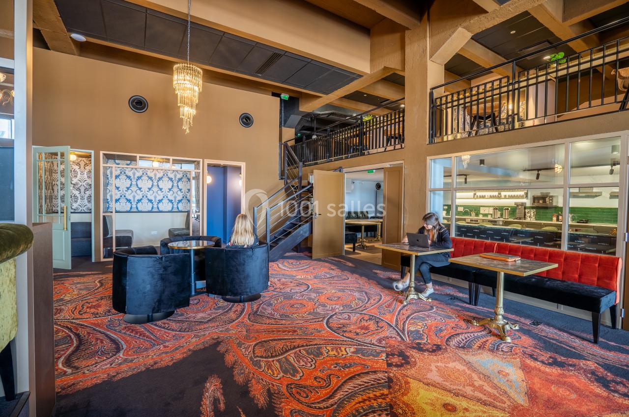 Intérieur d'un espace moderne avec moquette colorée, tables, fauteuils, escalier et personnes assises.