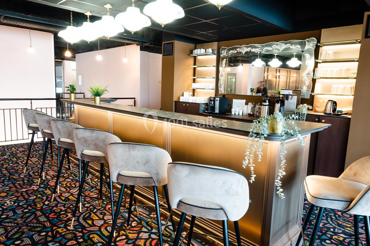 Comptoir de bar en bois clair avec tabourets, éclairé par des suspensions, sur un tapis coloré à motifs géométriques.