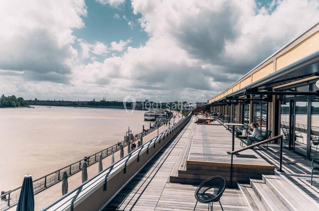 Terrasse en bois longeant un fleuve sous un ciel nuageux, avec des bâtiments et des personnes en arrière-plan.