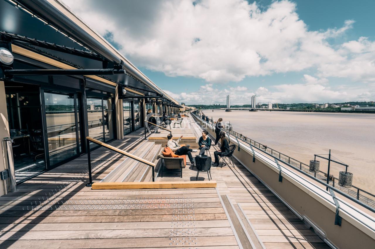 Terrasse en bois avec des tables et des chaises, surplombant une rivière sous un ciel partiellement nuageux.