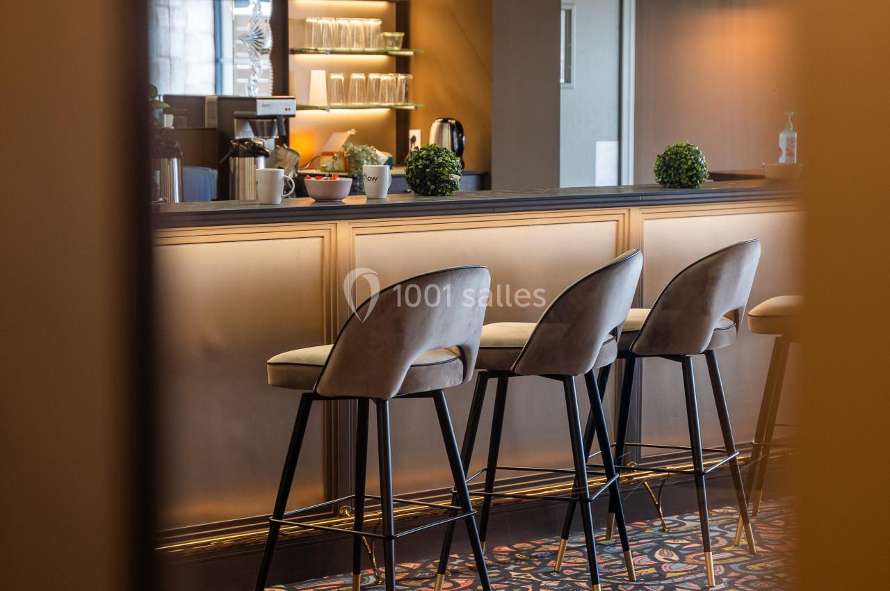 Bar élégant avec comptoir beige, tabourets rembourrés, suspensions lumineuses et sol à motifs colorés.