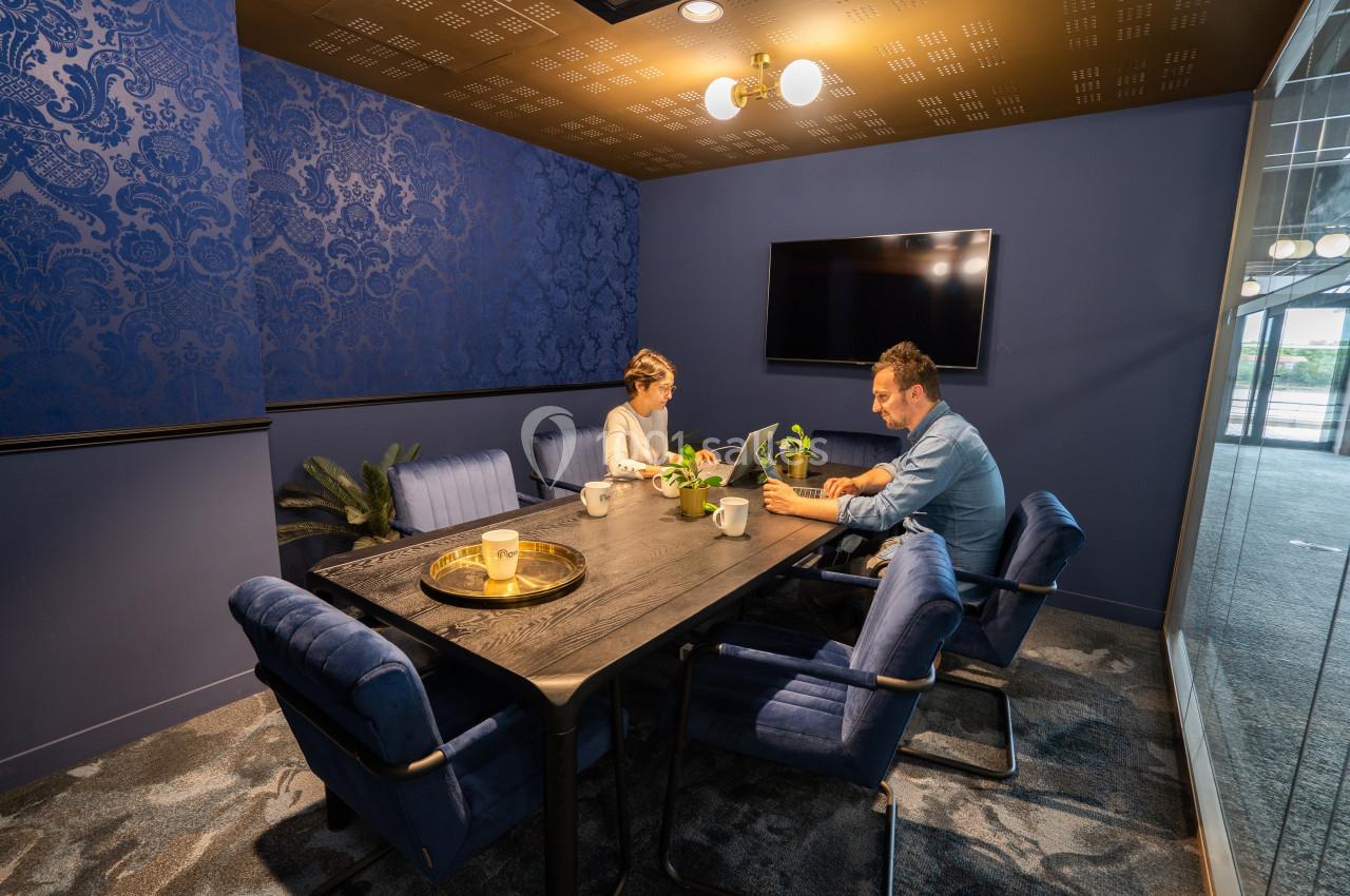 Deux personnes travaillent à une table dans une salle de réunion moderne avec murs bleus et éclairage chaleureux.