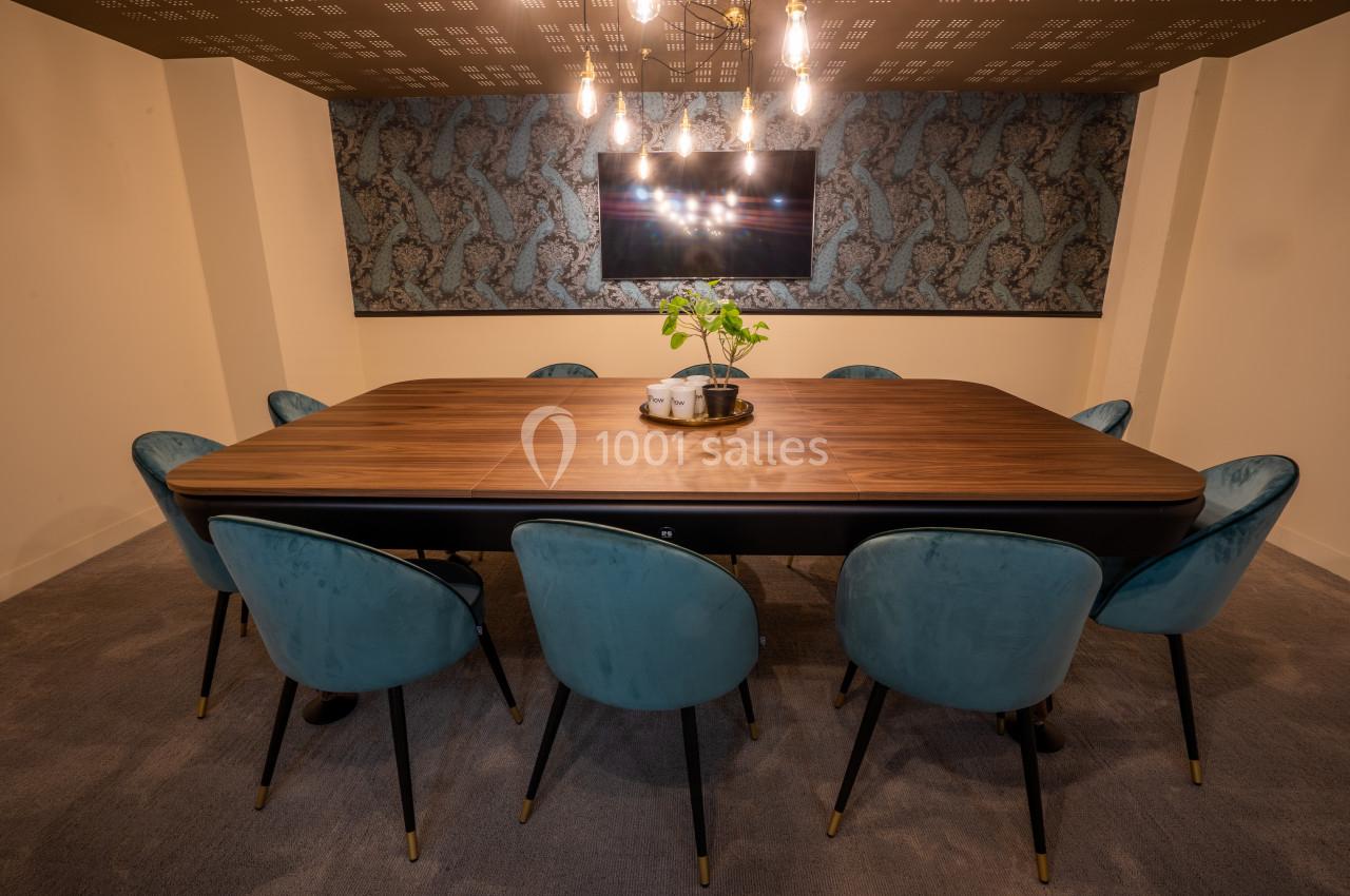 Salle de réunion avec table en bois, chaises bleues et éclairage suspendu, décorée d'un mur texturé et d'un écran.