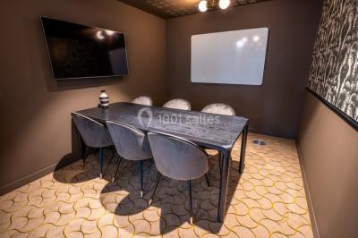 Salle de réunion avec table en bois foncé, six chaises grises, tableau blanc et écran mural.
