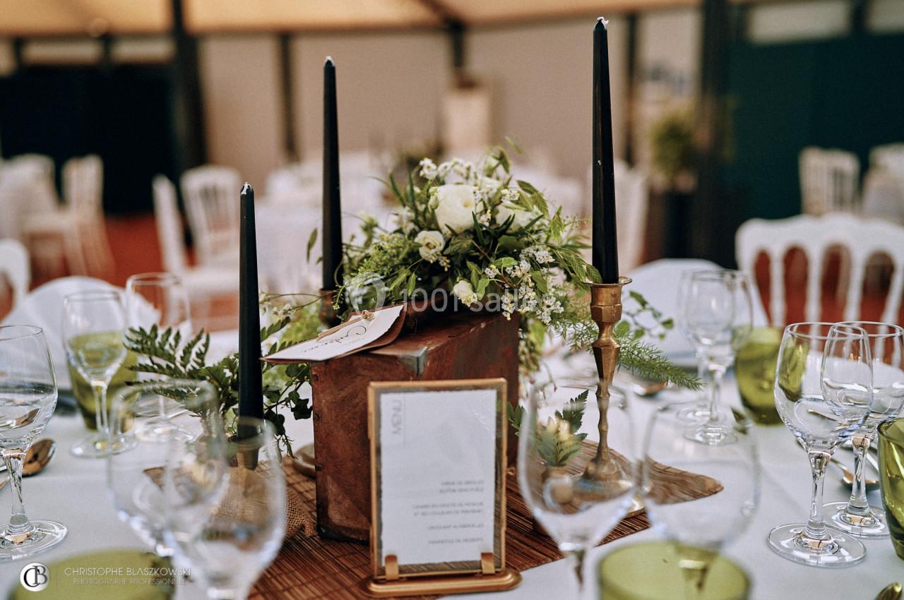 Centre de table avec fleurs blanches, bougies noires et éléments végétaux, entouré de verres et assiettes.