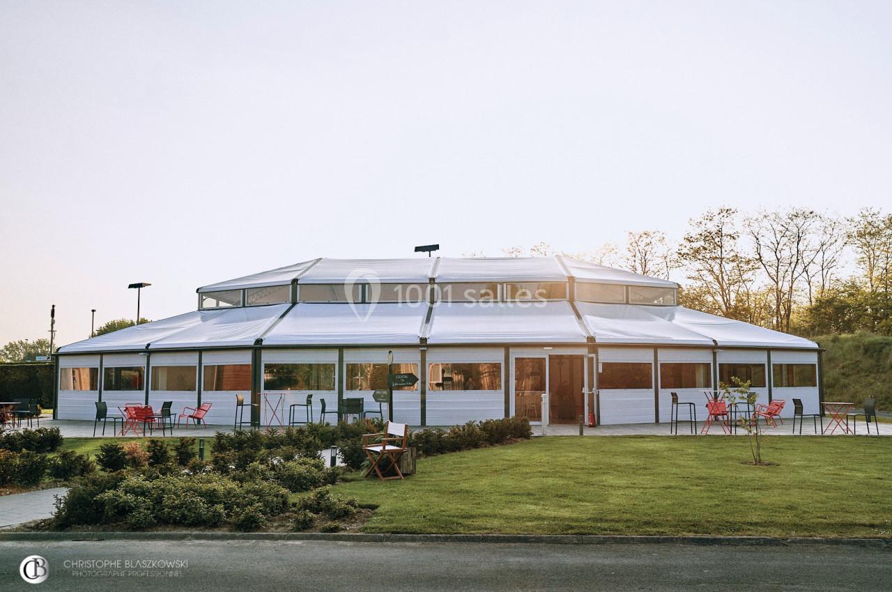 Bâtiment octogonal avec une structure blanche et des baies vitrées, entouré de verdure et de mobilier extérieur.