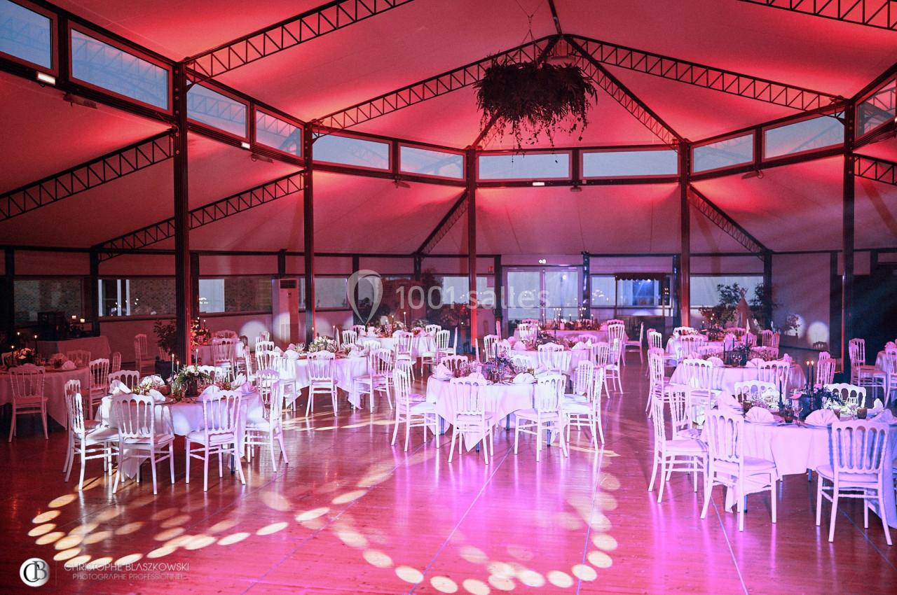 Salle de réception décorée avec des tables rondes, des chaises blanches et un éclairage rose tamisé.