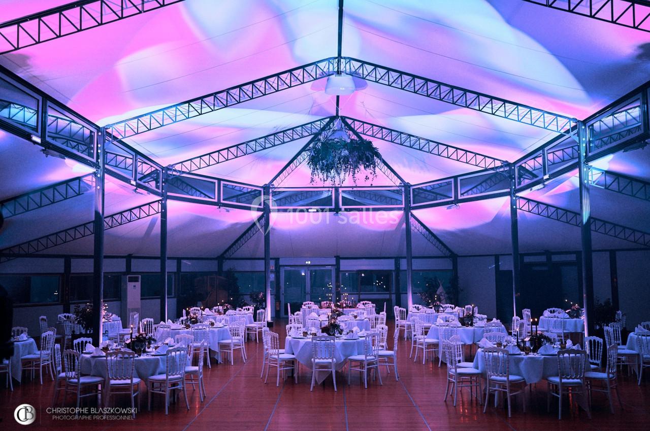 Salle de réception décorée avec des tables rondes, éclairage violet et bleu, et structure métallique apparente.