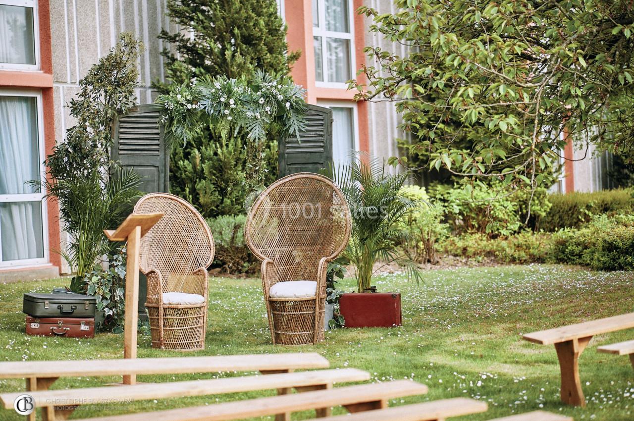Deux fauteuils en rotin placés devant une arche décorée de feuillage, dans un jardin avec des bancs en bois.