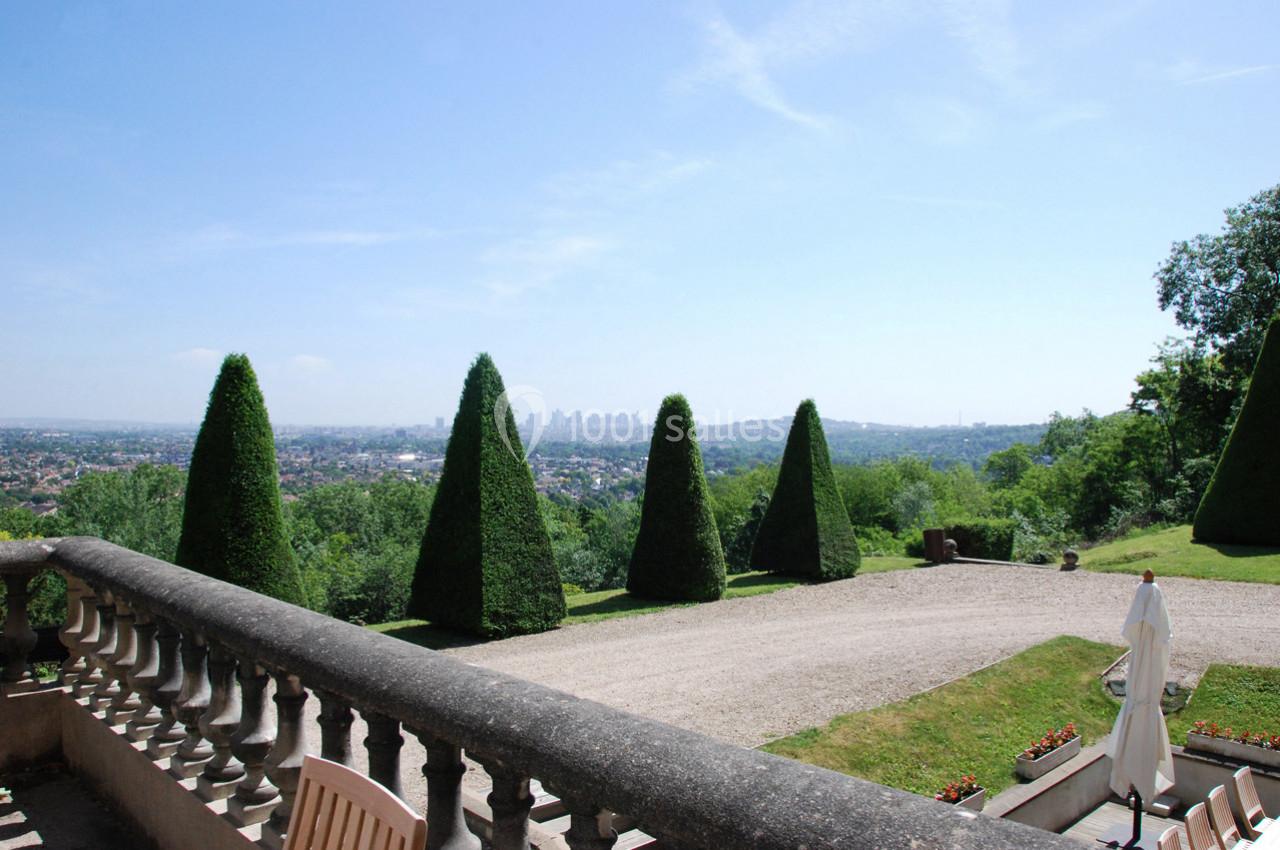 Vue depuis une terrasse avec balustrade sur un jardin, des arbres taillés en cônes et un paysage urbain à l'horizon.