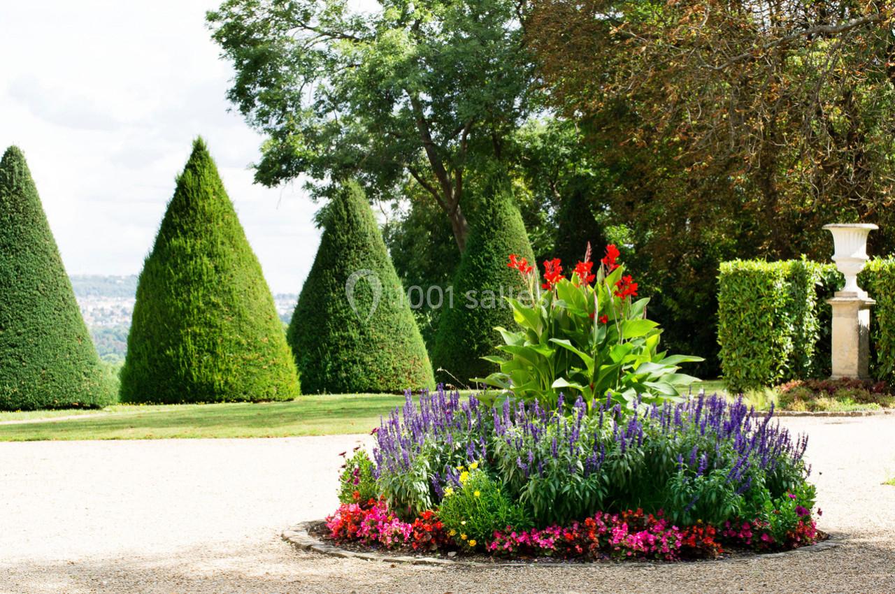 Massif fleuri coloré entouré de pelouse, avec des arbres taillés en cônes et un paysage verdoyant en arrière-plan.