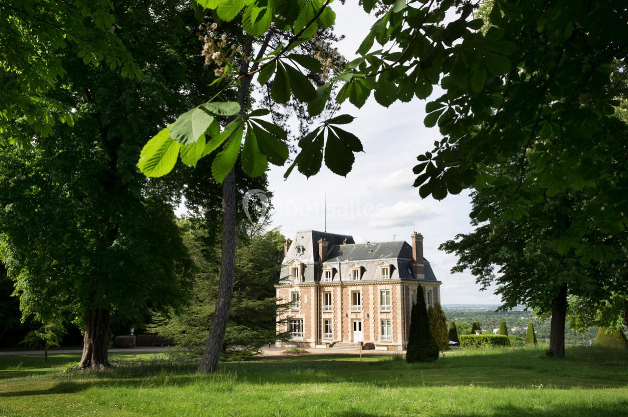 Manoir en pierre entouré d'arbres et d'une pelouse, sous un ciel partiellement nuageux.