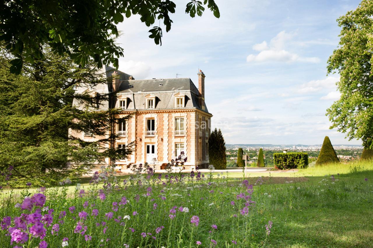 Manoir en pierre entouré de verdure, avec un jardin fleuri au premier plan et un ciel dégagé en arrière-plan.