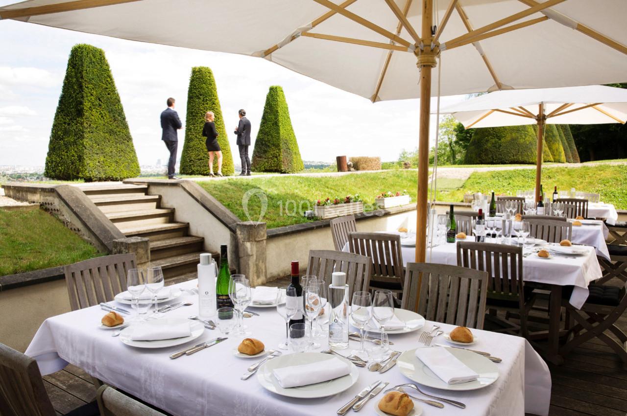 Terrasse d'un restaurant avec tables dressées, vue sur un jardin et trois personnes debout près de haies taillées.