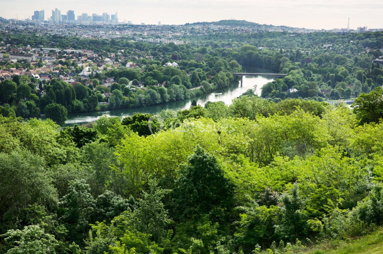 Vue d'une vallée verdoyante avec une rivière sinueuse, des collines boisées et des bâtiments en arrière-plan.