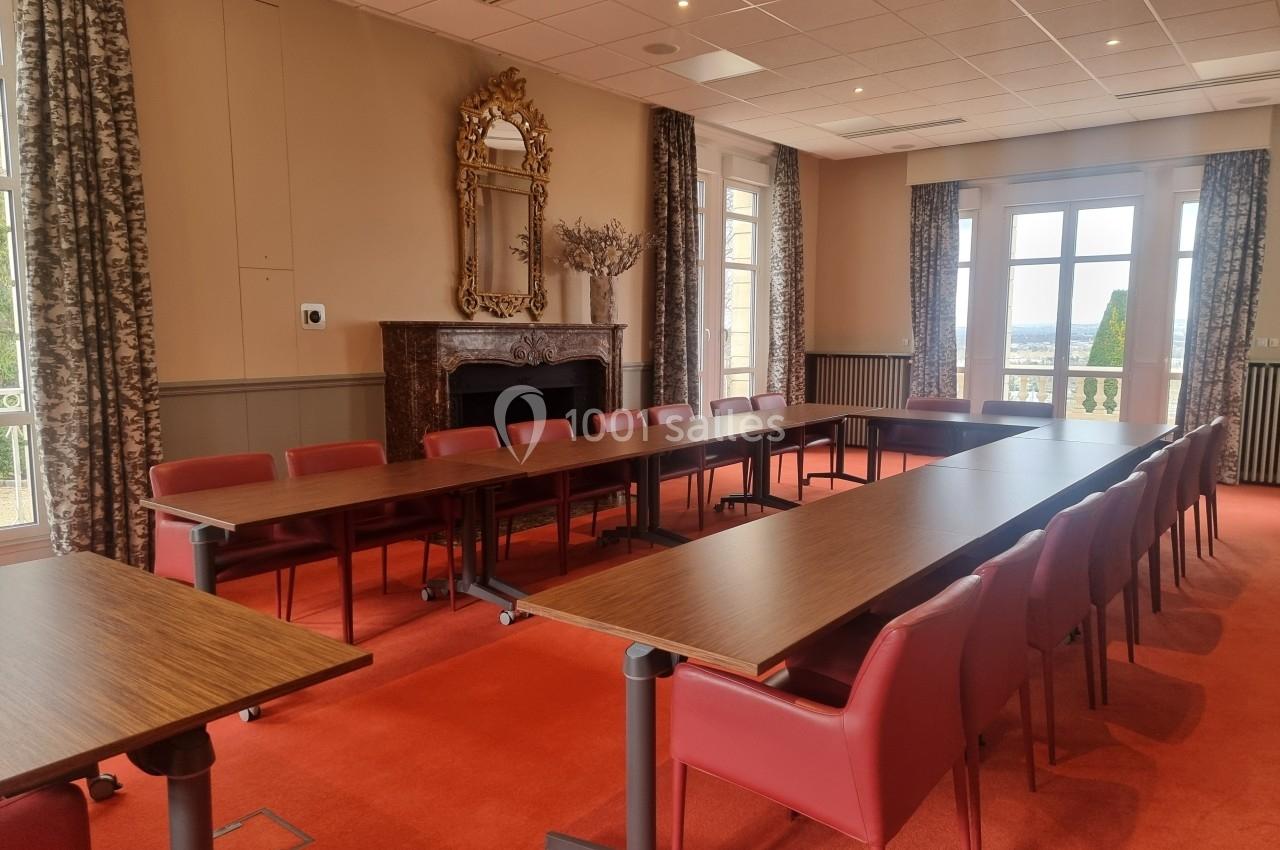 Salle de réunion lumineuse avec tables en bois, chaises rouges, cheminée, miroir et grandes fenêtres donnant sur l'extérieur.