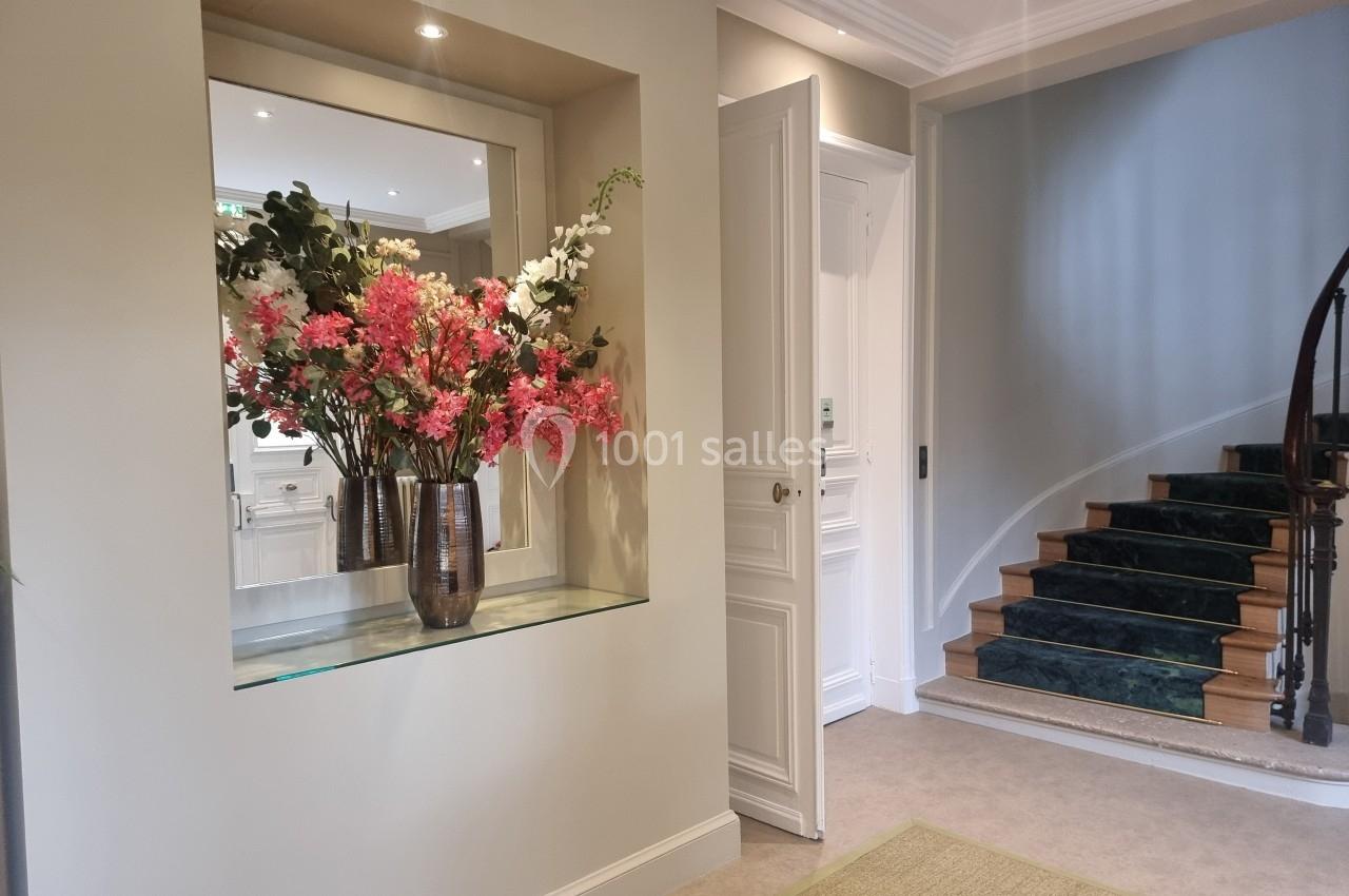Un bouquet de fleurs colorées dans un vase métallique, posé sur une étagère devant un miroir dans un hall lumineux.