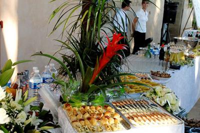 Tables rondes dressées avec nappes blanches, serviettes roses et verres, disposées en extérieur sous des parasols.