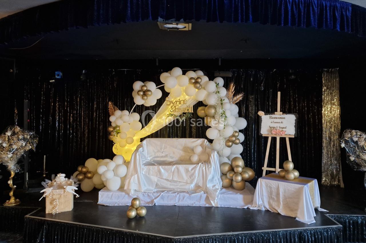 Décor de scène avec arche en ballons blancs et dorés, fauteuil central, et panneau de bienvenue sur chevalet.