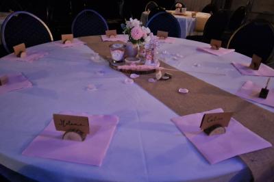 Tables rondes dressées avec nappes blanches, serviettes roses et verres, disposées en extérieur sous des parasols.