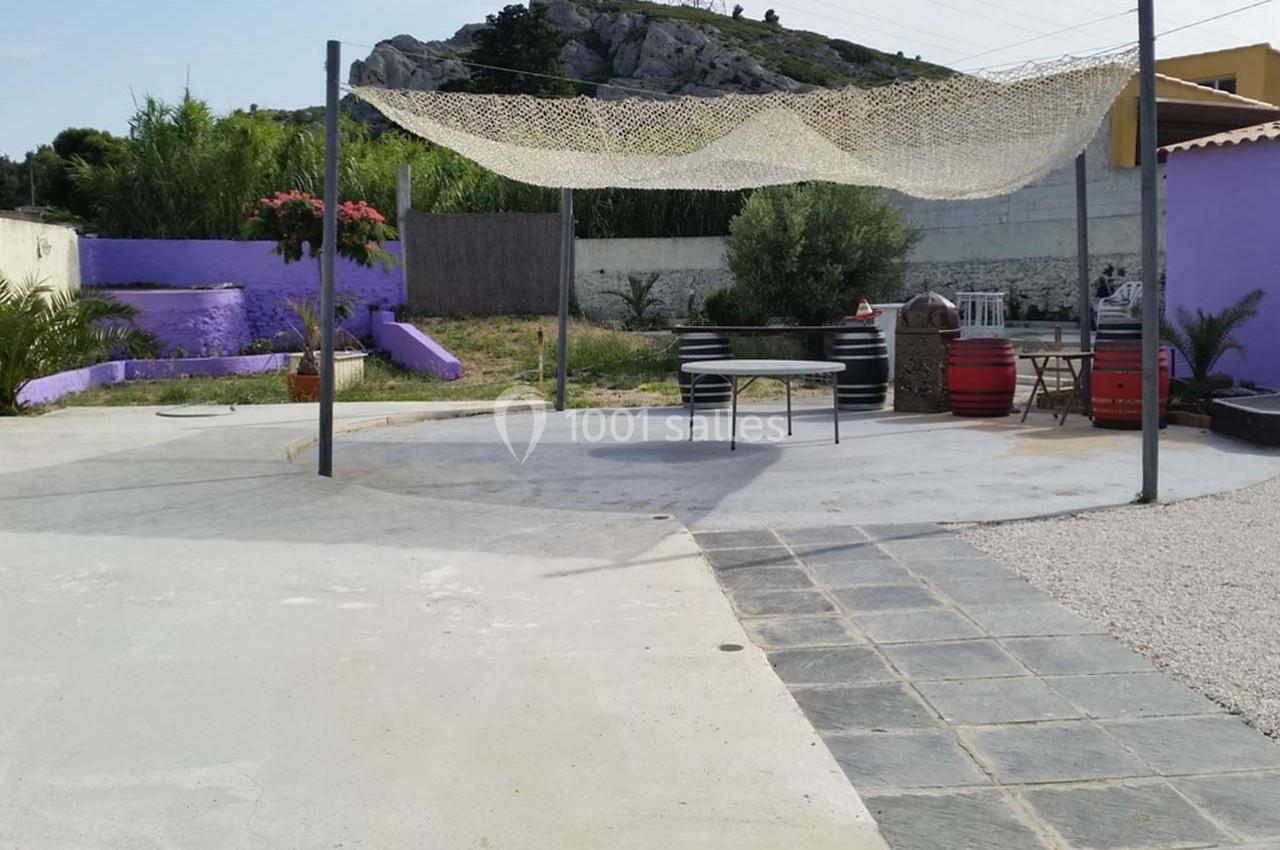 Terrasse extérieure avec voile d'ombrage, table, chaises, murs violets, végétation et colline en arrière-plan.