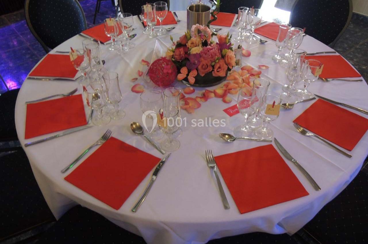Table ronde dressée avec nappes blanches, serviettes rouges, couverts, verres et centre de table floral coloré.