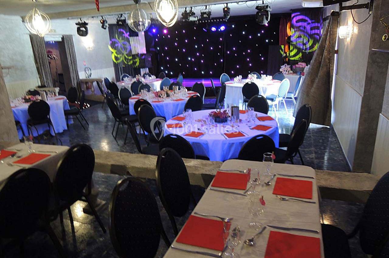 Salle de réception décorée avec des tables rondes et rectangulaires, nappes blanches et serviettes rouges, scène en arrière…