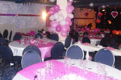 Tables rondes dressées avec nappes blanches, serviettes roses et verres, disposées en extérieur sous des parasols.