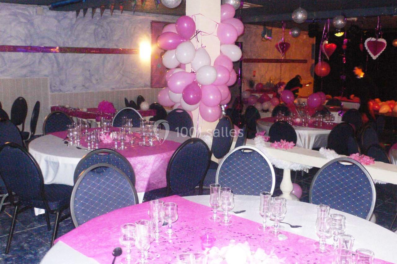 Salle décorée pour une fête avec des ballons roses et blancs, nappes roses, tables dressées et chaises bleues.