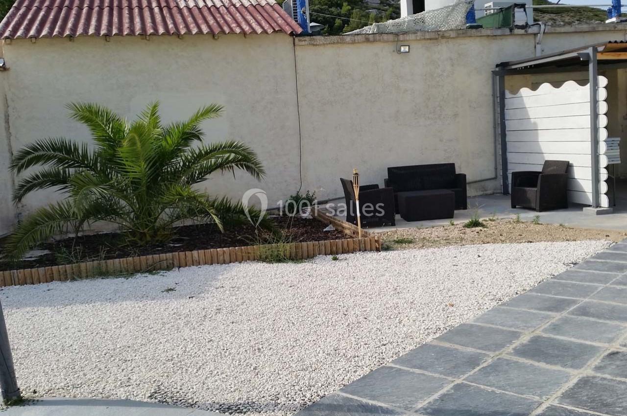 Cour extérieure avec gravier blanc, petit palmier, mobilier de jardin en rotin et mur beige en arrière-plan.