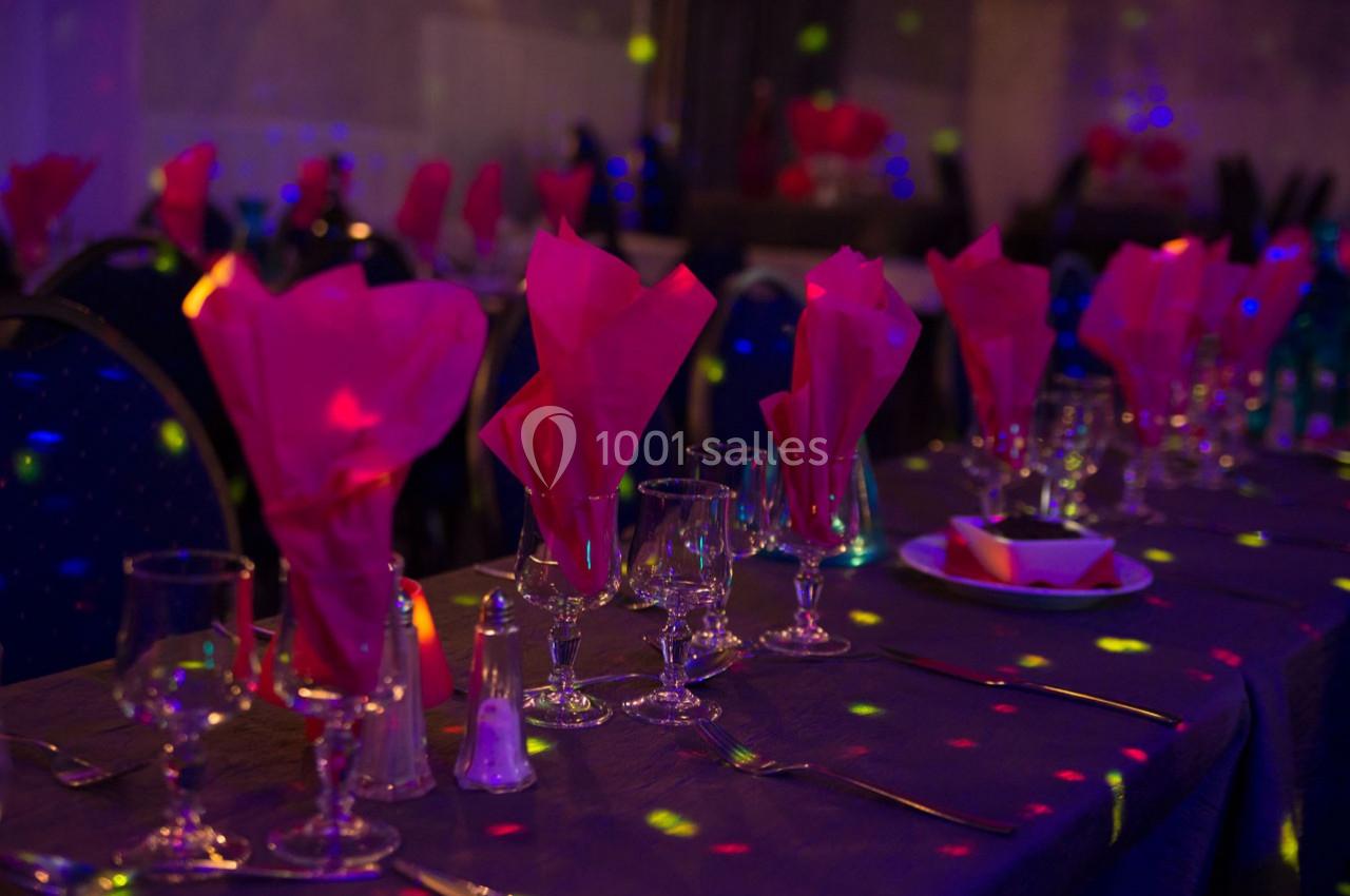 Table dressée avec nappes sombres, serviettes roses pliées, verres et couverts, éclairée par des lumières colorées.