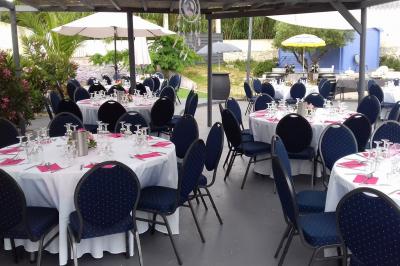 Tables rondes dressées avec nappes blanches, serviettes roses et verres, disposées en extérieur sous des parasols.