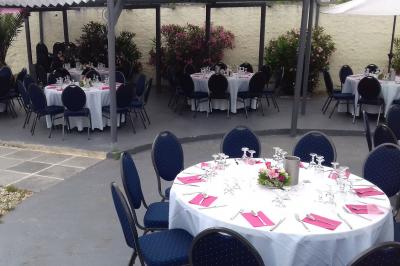 Tables rondes dressées avec nappes blanches, serviettes roses et verres, disposées en extérieur sous des parasols.
