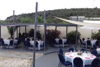 Tables rondes dressées avec nappes blanches, serviettes roses et verres, disposées en extérieur sous des parasols.